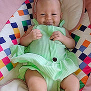 Athena is registered to the contest to win money with this photo: baby, smiling, child, green_outfit, colorful_cushion, pink_border, infant, cute, happy, portrait, indoors, seated, soft_lighting, comfortable, playful, young_child, relaxed, human, small_hands, fabric