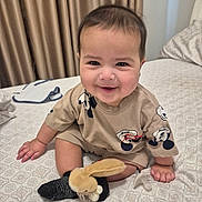 Massimo is registered to the contest to win money with this photo: baby, child, smiling, indoor, bed, blanket, toy, plush, mickey_mouse, clothing, socks, face, person, curtains, pillow, cute, happy, seated, young_child, home