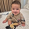 baby, child, smiling, indoor, bed, blanket, toy, plush, mickey_mouse, clothing, socks, face, person, curtains, pillow, cute, happy, seated, young_child, home