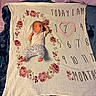 baby, infant, child, sleeping, pacifier, portrait, milestone_blanket, floral, heart, numbers, handwritten_text, bed, blanket, pillows, bedroom, indoor, lying_down, onesie, patterned_pants, bare_feet