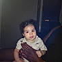 baby, child, curly_hair, onesie, hand, adult, indoor, person, sitting, cute, infant, holding, dark_skin, expression, portrait, looking, eyes, small, room, door