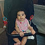 baby, black_stroller, carriage, child, clothing, curly_hair, cute, expression, face, feet, floor, hands, indoor, infant, person, pink_bib, relaxed, sitting, stroller, toy