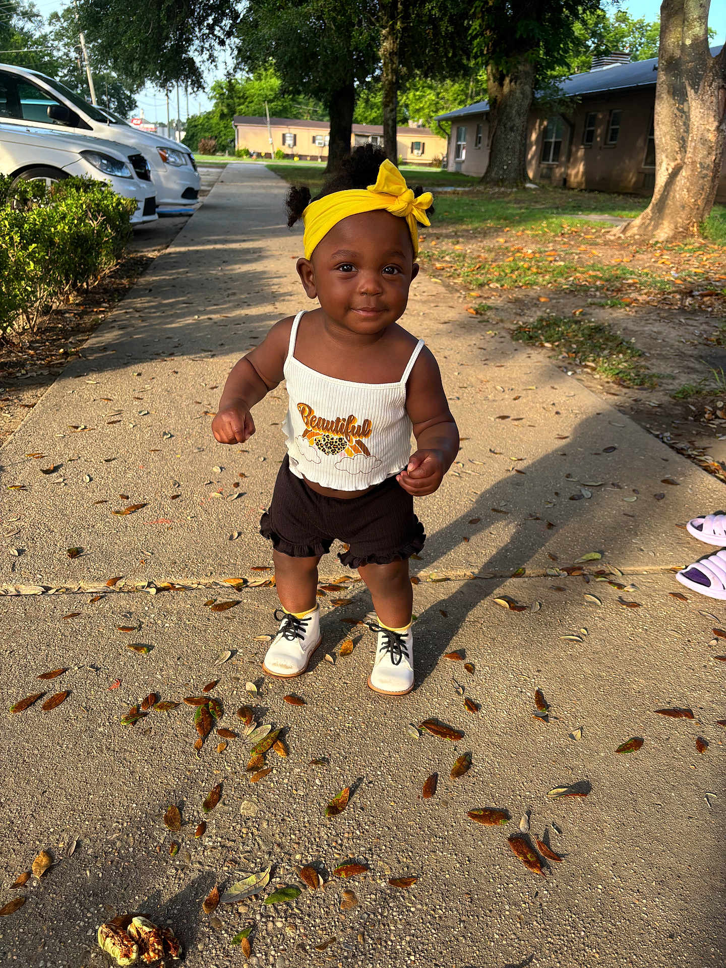 Nova is registered to the contest to win money with this photo: toddler, child, outdoor, sidewalk, yellow_headband, white_shoes, black_shorts, sunlight, shadow, tree, leaves, smile, cute, person, summer, casual_clothing, park, happy, walking, daytime