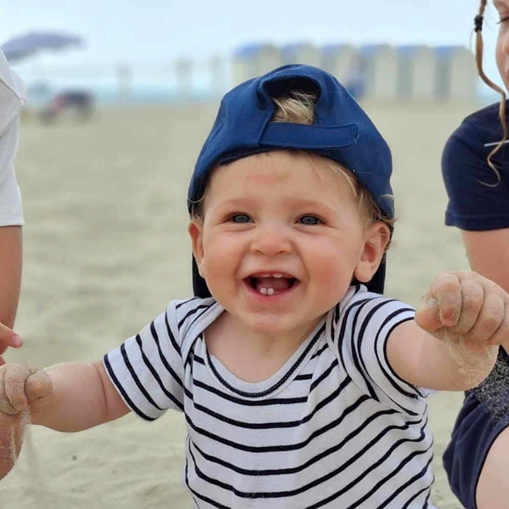 Naé a rejoint le concours — aidez-le/la à gagner de superbes lots ! baby, beach, cap, casual_clothing, child, cute, daytime, fun, happy, joy, outdoor, person, play, sand, sitting, smiling, striped_shirt, summer, sunny, toddler