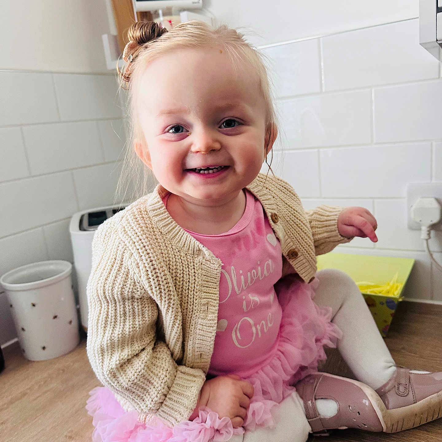 Olivia Harper is registered to the contest to win money with this photo: child, cute, face, floor, hair_bun, happy, indoors, kitchen, knitted_cardigan, light, person, pink_dress, shoes, sitting, smiling, toddler, wall_tiles, white_tights, wooden_floor, young