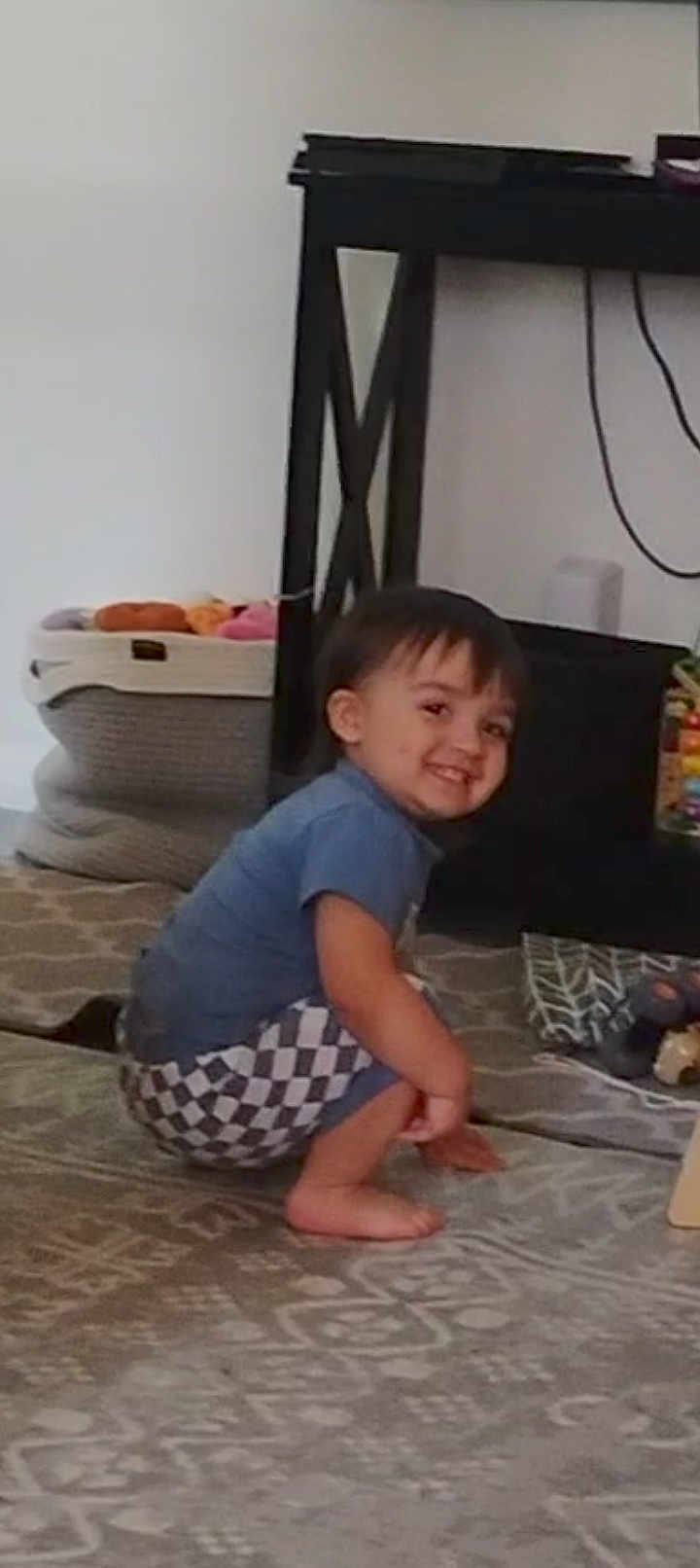 Leo is registered to the contest to win money with this photo: child, toddler, smiling, indoor, carpet, toy, basket, furniture, table, shirt, shorts, person, hair, floor, home, cute, happy, playful, barefoot, living_room