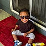 child, toddler, sunglasses, toy, red_rug, door, sneakers, blue, white, black_shirt, gray_pants, outdoor, concrete_floor, smiling, sitting, person, play, fun, casual, young