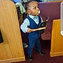 toddler, child, pacifier, blue_suit, bow_tie, black_shoes, church, pew, carpet, wooden_pew, person, indoor, holding_object, standing, curious, people_in_background, formal_wear, young_child, hat, stick