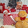 baby, background_tree, blanket, child, christmas, cute, festive_clothing, floor_rug, gift_box, gingerbread_man, happy, holiday_decor, holiday_theme, indoors, ornaments, plush_toy, red_hat, smiling, toy, wrapped_present