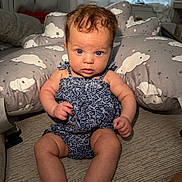 Evelyn joined the competition — help win amazing prizes! baby, child, infant, blue_eyes, curly_hair, romper, cushion, cloud_pattern, indoor, soft_lighting, sitting, cute, curious, skin, person, furniture, room, floor, support, portrait