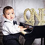 toddler, child, piano, miniature_piano, bow_tie, white_shirt, black_pants, celebration, birthday, number_one, gold_glitter, music, indoors, portrait, young_child, hands_on_piano, expression, face, cute, formal_wear