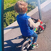 William participe au concours pour gagner de l'argent avec cette photo : active, back_view, balance_bike, bike, blue_vest, casual_clothing, child, daytime, footwear, grass, jeans, outdoor, pavement, person, play, recreation, shadow, sneakers, sunlight, toddlers