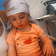 Judith participe au concours pour gagner de l'argent avec cette photo : child, toddler, baby, bandana, headscarf, onesie, orange_clothing, sitting, hand, face, eyes_down, long_eyelashes, blanket, floral_pattern, wall, outdoor, sunlight, grill, adult_hand, tattoo