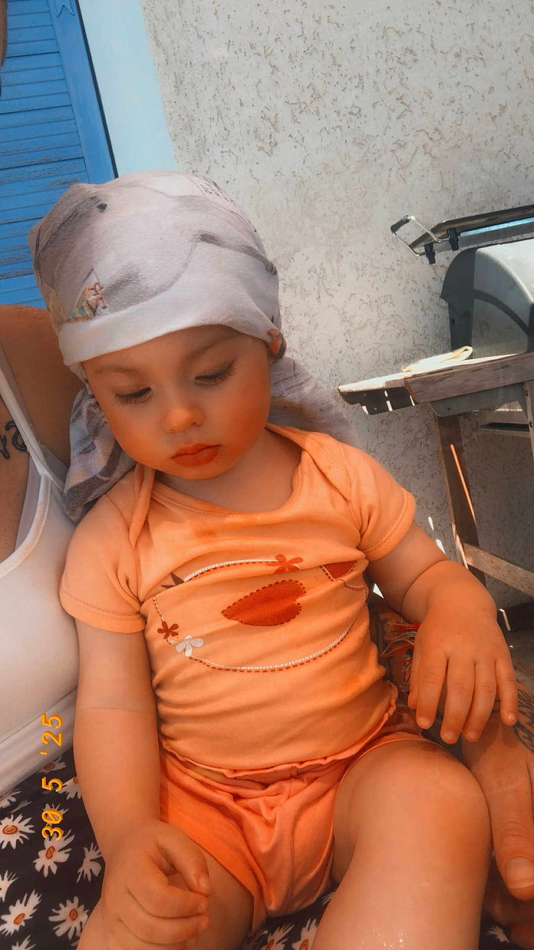 Judith participe au concours pour gagner de l'argent avec cette photo : child, toddler, baby, bandana, headscarf, onesie, orange_clothing, sitting, hand, face, eyes_down, long_eyelashes, blanket, floral_pattern, wall, outdoor, sunlight, grill, adult_hand, tattoo