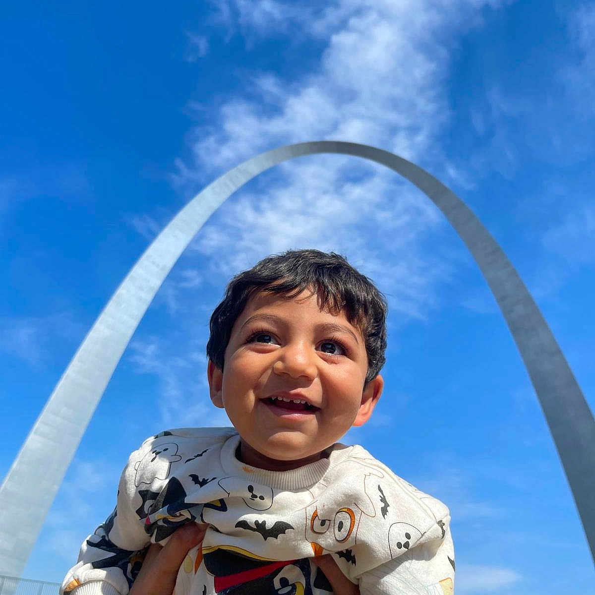Subh is registered to the contest to win money with this photo: child, boy, smiling, happy, outdoor, blue_sky, clouds, arch, landmark, hands, sweater, cartoon_print, daylight, portrait, person, playful, holding, architecture, tourist_spot, sky