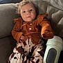 baby, infant, child, couch, clothing, patterned_pants, curly_hair, sippy_cup, indoor, face, person, relaxing, seated, cozy, soft_lighting, home, furniture, cute, looking_at_camera, casual