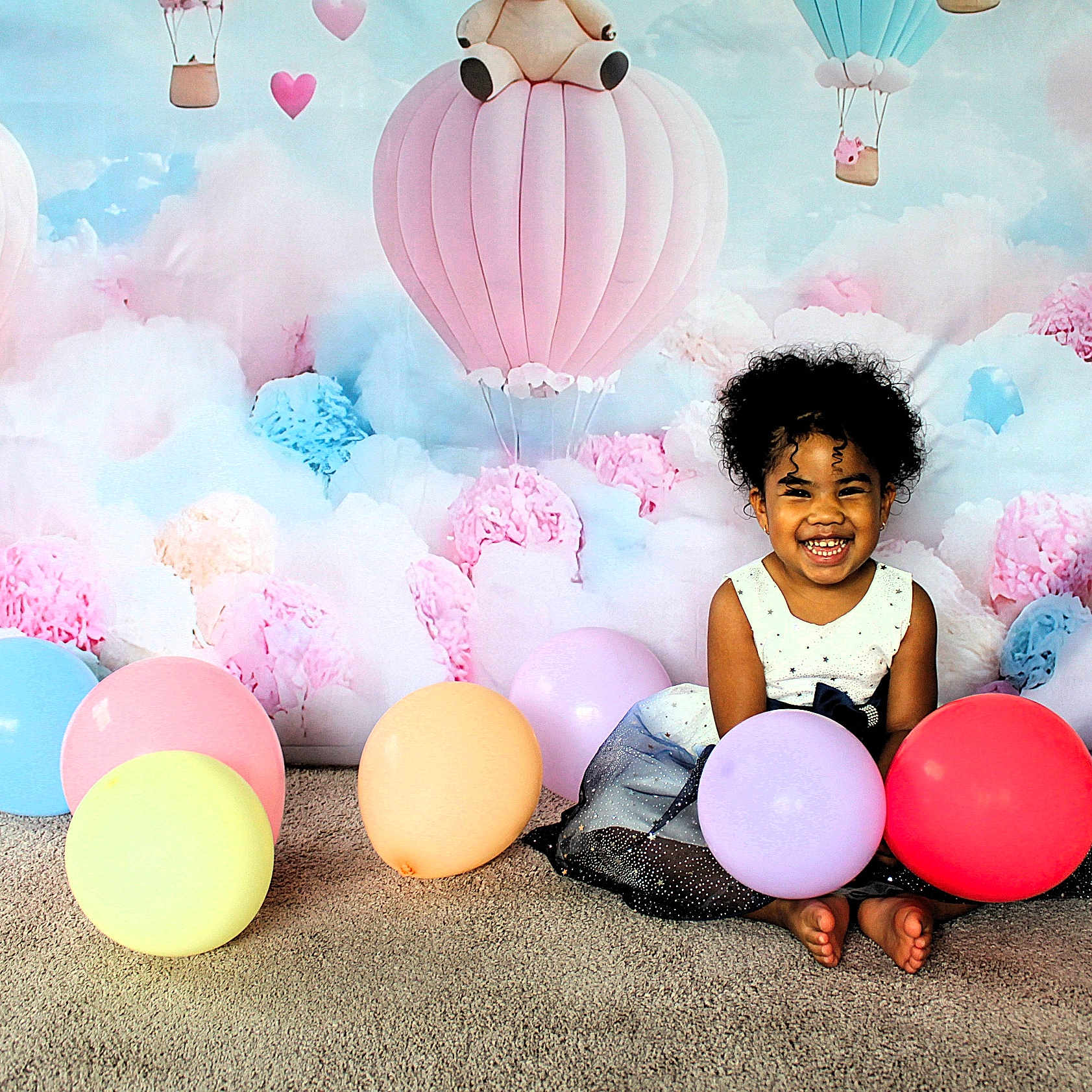 Kataléya participe au concours pour gagner de l'argent avec cette photo : background, balloons, barefoot, carpet, celebration, child, colorful, curly_hair, cute, decor, dress, happy, hot_air_balloon, indoor, pastel_colors, playful, portrait, sitting, smile, toddler