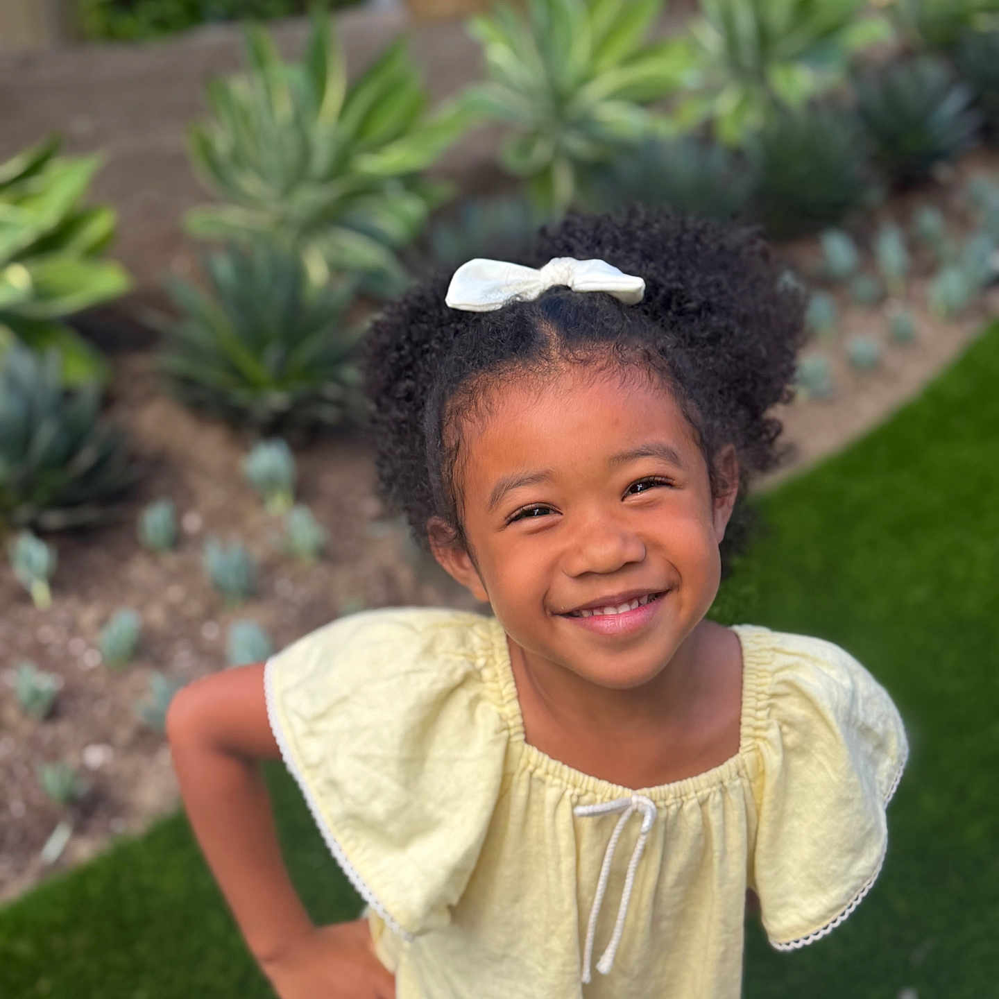 Kamilla is registered to the contest to win money with this photo: child, girl, smiling, curly_hair, bow, yellow_top, blue_jeans, outdoor, garden, grass, plants, portrait, happy, casual_clothing, sunlight, greenery, person, young, cute, playful