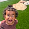 child, toddler, smiling, curly_hair, outdoor, grass, rocks, happy, portrait, person, casual_clothing, greenery, cute, young, natural_light, fun, joy, face, playful, boy