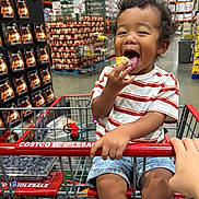 Colin is registered to the contest to win money with this photo: toddler, child, shopping_cart, costco, food, warehouse, shelves, snack, curly_hair, striped_shirt, happy, smiling, indoor, supermarket, retail, person, hand, clothing, blue_shorts, grocery
