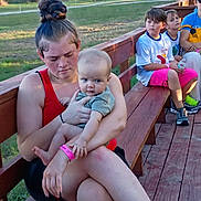 Riley is registered to the contest to win money with this photo: baby, child, woman, bench, outdoor, greenery, grass, wood, people, casual_clothing, wristband, summer, park, sitting, holding, curious, sunlight, daytime, children, relaxed