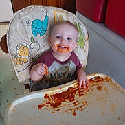 Benjamin is registered to the contest to win money with this photo: toddler, child, high_chair, messy, food, tomato_sauce, smile, happy, face, hand, kitchen, cabinet, floor, clothing, baby, person, indoor, feeding, plate, fun