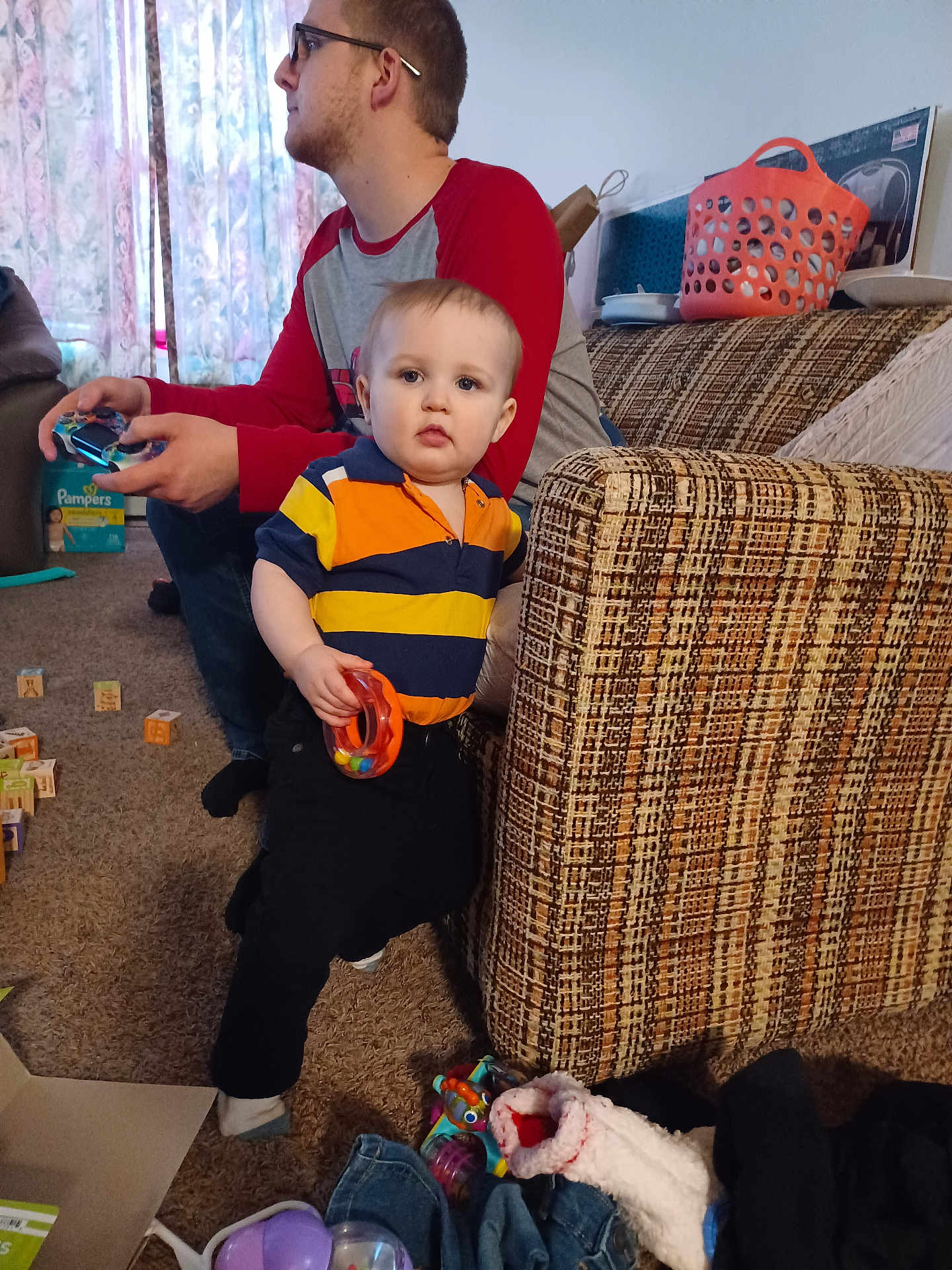 Benjamin joined the competition — help win amazing prizes! toddler, man, video_game_controller, couch, toy, carpet, window, curtains, box, clothing, glasses, indoor, person, child, living_room, furniture, sock, basket, jeans, plush_toy