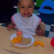 Amirah is registered to the contest to win money with this photo: toddler, child, smiling, braids, beads, bib, table, plate, cupcake, crackers, cheese_puffs, food, indoor, chair, green_wall, blue_chair, happy, person, portrait, kid