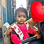 child, toddler, pink_clothing, pony_tail, stroller, safety_bar, adult, heart_emoji, bus_interior, window, hand, seat, curious, indoor, person, transportation, public_transport, face, holding, casual