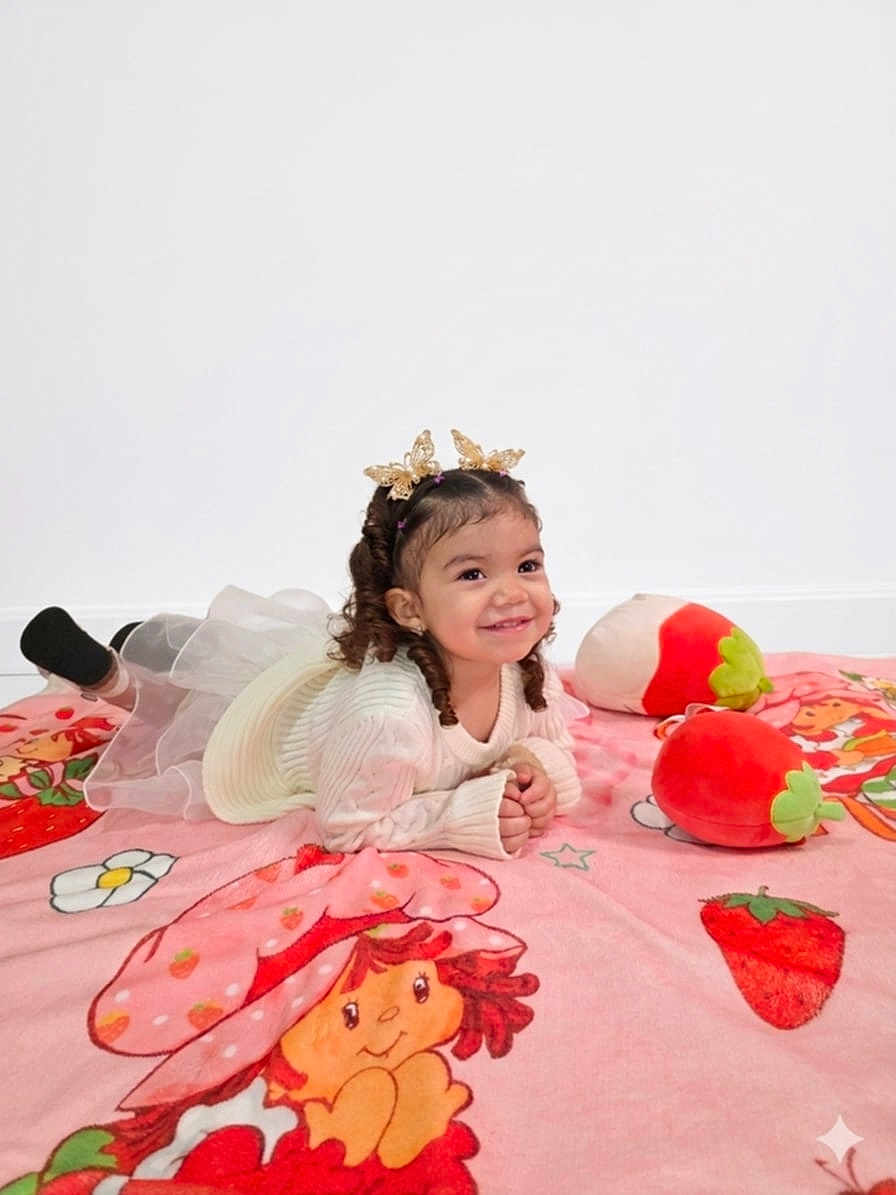 Kler is registered to the contest to win money with this photo: child, toddler, girl, smiling, blanket, plush_toy, strawberry, pink, headband, crown, dress, curly_hair, lying_down, toy, socks, rug, happy, portrait, studio, playful