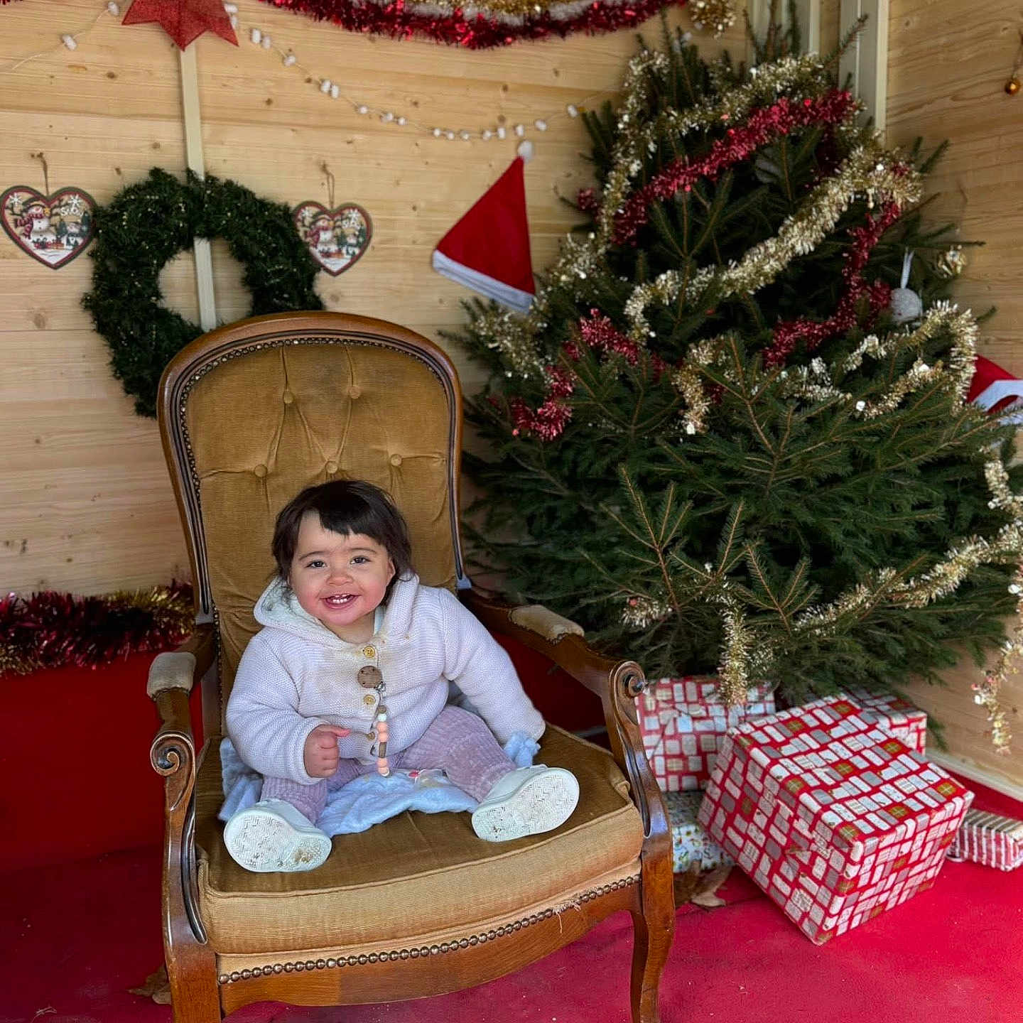 Lina a rejoint le concours — aidez-le/la à gagner de superbes lots ! child, toddler, smiling, chair, armchair, christmas_tree, christmas_decorations, presents, gift_wrap, red_carpet, wooden_wall, garland, red_bow, holiday, festive, indoors, happy, cozy, person, seasonal