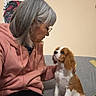 woman, dog, cavalier_king_charles_spaniel, pet, companion, sitting, couch, indoor, portrait, glasses, gray_hair, pink_hoodie, white_and_brown_fur, floppy_ears, looking_at_each_other, home, wall, lamp, blanket, cozy
