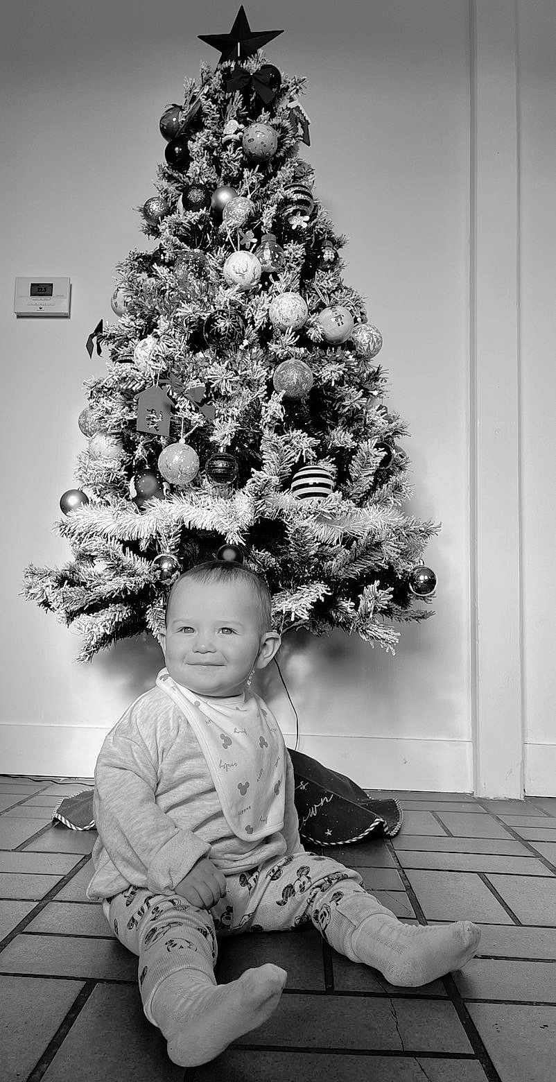 Marius participe au concours pour gagner de l'argent avec cette photo : baby, child, christmas_tree, holiday, pajamas, bib, smiling, indoor, floor, decorations, ornaments, pine, star, sitting, tiled_floor, person, festive, monochrome, cute, young_child