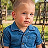 child, toddler, boy, portrait, face, eyes, short_hair, denim_shirt, clothing, sitting, swing, playground, outdoor, park, sunlight, trees, grass, playground_equipment, close_up, photography