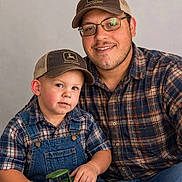 Parker is registered to the contest to win money with this photo: child, toddler, adult, father, portrait, smiling, family, indoors, plaid_shirt, baseball_cap, john_deere, overalls, toy_tractor, tractor, father_son, sitting, eyeglasses, beard, casual_clothing, photograph