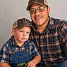 child, toddler, adult, father, portrait, smiling, family, indoors, plaid_shirt, baseball_cap, john_deere, overalls, toy_tractor, tractor, father_son, sitting, eyeglasses, beard, casual_clothing, photograph