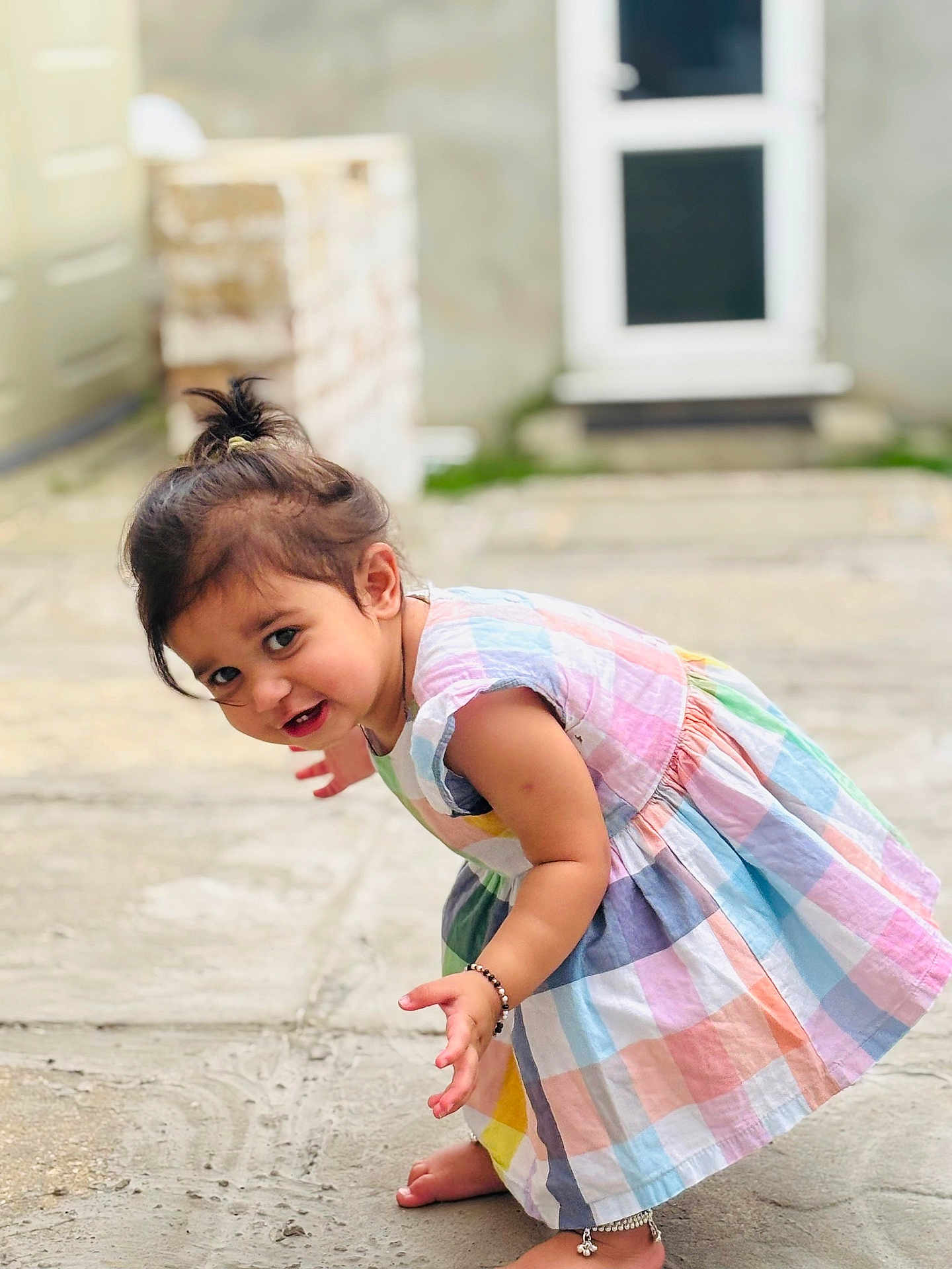 Risha joined the competition — help win amazing prizes! toddler, child, dress, colorful, outdoor, curious, barefoot, bracelet, anklet, smiling, candid, portrait, playful, young_child, person, hair_tied, pavement, door, background_blur, sunlight