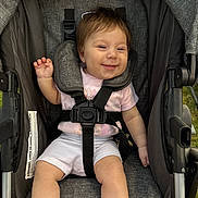 Lainey is registered to the contest to win money with this photo: baby, child, stroller, smile, happy, pink_shirt, white_shorts, sitting, seatbelt, outdoor, grass, cute, infant, person, young_child, portrait, one_person, clothing, hand, foot