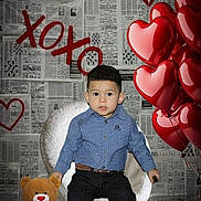 Liam is registered to the contest to win money with this photo: child, boy, teddy_bear, heart_balloon, balloon, newspaper_background, xoxo, red_heart, blue_shirt, black_pants, sneakers, white_chair, indoor, cute, portrait, smiling, love_theme, decor, seated, playful