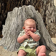 Johnathan is registered to the contest to win money with this photo: baby, child, sitting, tree_stump, sand, outdoor, sunlight, clothing, cute, happy, hands, smile, nature, skin, barefoot, infant, shorts, patterned_shirt, daylight, relaxing