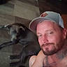 animal, beard, cap, carpet, casual, chain, dog, floor, furniture, human, indoor, man, pet, portrait, relaxed, room, rug, selfie, tattoo, wooden_floor