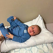 Erik is registered to the contest to win money with this photo: baby, infant, blue_onesie, pillow, bed, blanket, person, child, smiling, happy, hand, foot, wall, bedroom, cushion, sleepwear, skin, head, young_child, indoor