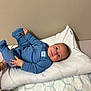 baby, infant, blue_onesie, pillow, bed, blanket, person, child, smiling, happy, hand, foot, wall, bedroom, cushion, sleepwear, skin, head, young_child, indoor