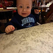 Erik is registered to the contest to win money with this photo: toddler, child, table, restaurant, marble_table, clothing, sweatshirt, jordan, face, person, indoor, people, seated, fist, curious, wide_eyes, young_child, casual_wear, background_people, dining