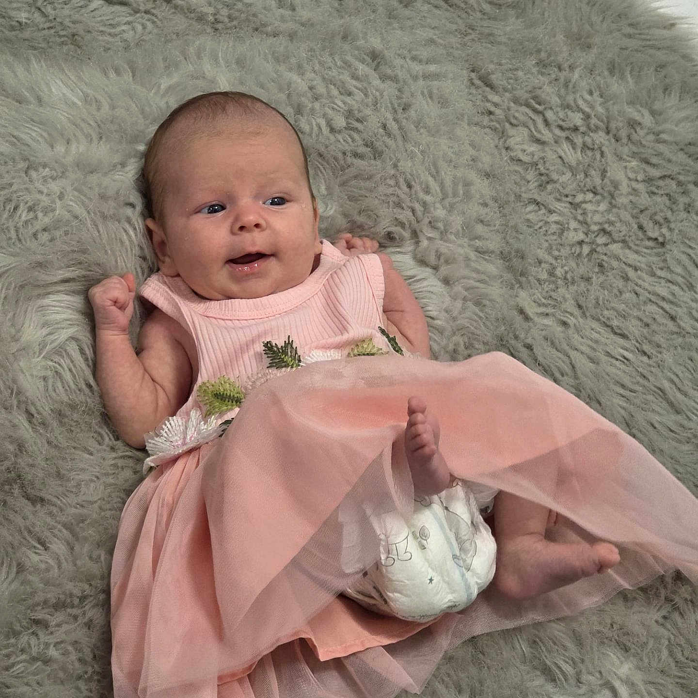 Astrid is registered to the contest to win money with this photo: baby, barefoot, child, clothing, cute, diaper, face, fluffy_rug, happy, head, indoors, infant, lying_down, newborn, pink_dress, playful, skin, small, smiling, soft_texture