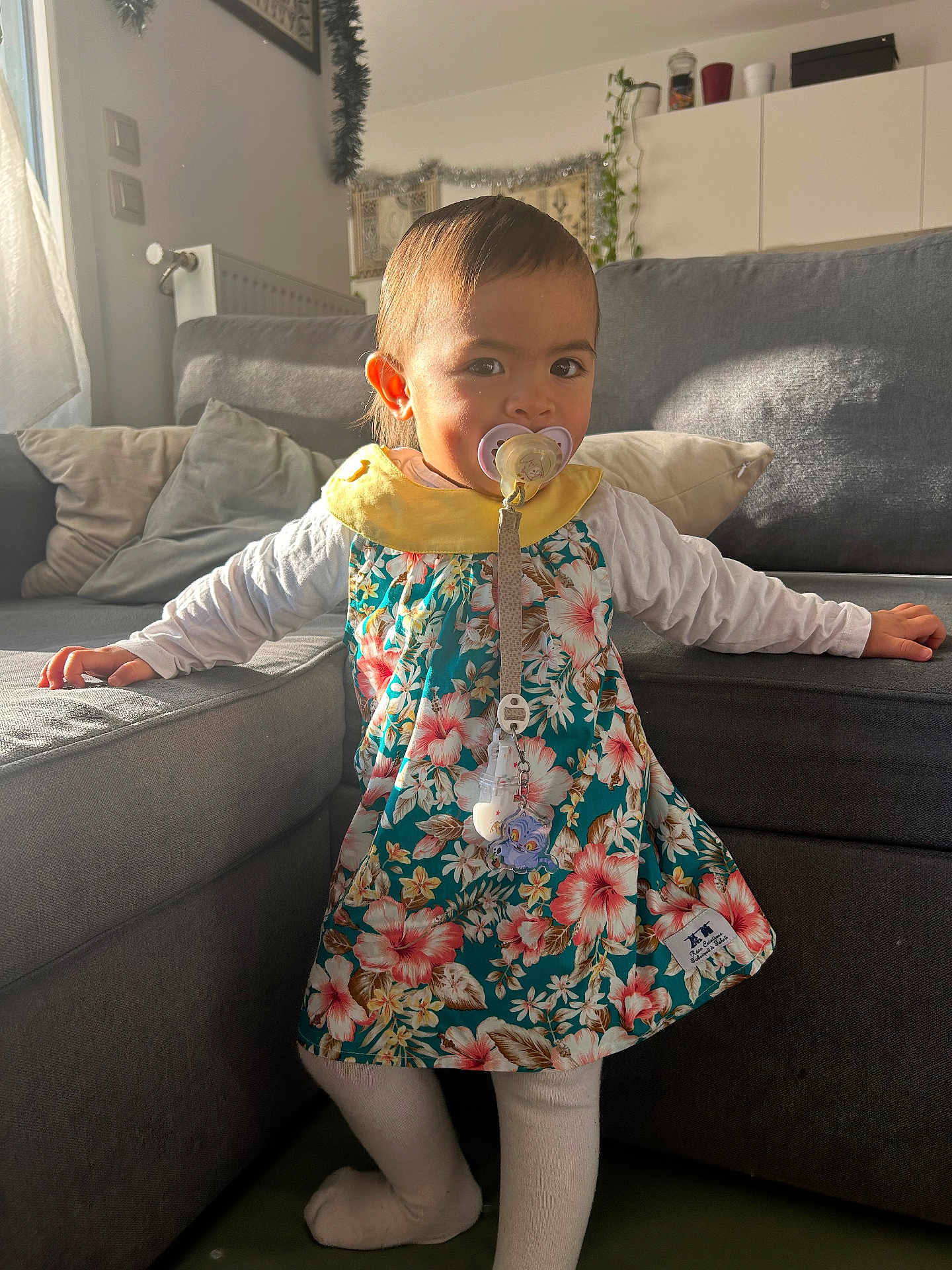Owena a rejoint le concours — aidez-le/la à gagner de superbes lots ! child, toddler, pacifier, floral_dress, dress, sofa, couch, pillow, living_room, sunlight, shadow, standing, home_interior, cute, face, long_sleeves, tights, pacifier_clip, curtain, plant