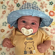 Maple is registered to the contest to win money with this photo: baby, pacifier, hat, blue_hat, yellow_onesie, bear_design, flower_pattern, blanket, infant, cute, lying_down, clothing, child, portrait, indoors, soft_texture, hand, face, wide_eyes, headwear