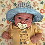 baby, pacifier, hat, blue_hat, yellow_onesie, bear_design, flower_pattern, blanket, infant, cute, lying_down, clothing, child, portrait, indoors, soft_texture, hand, face, wide_eyes, headwear