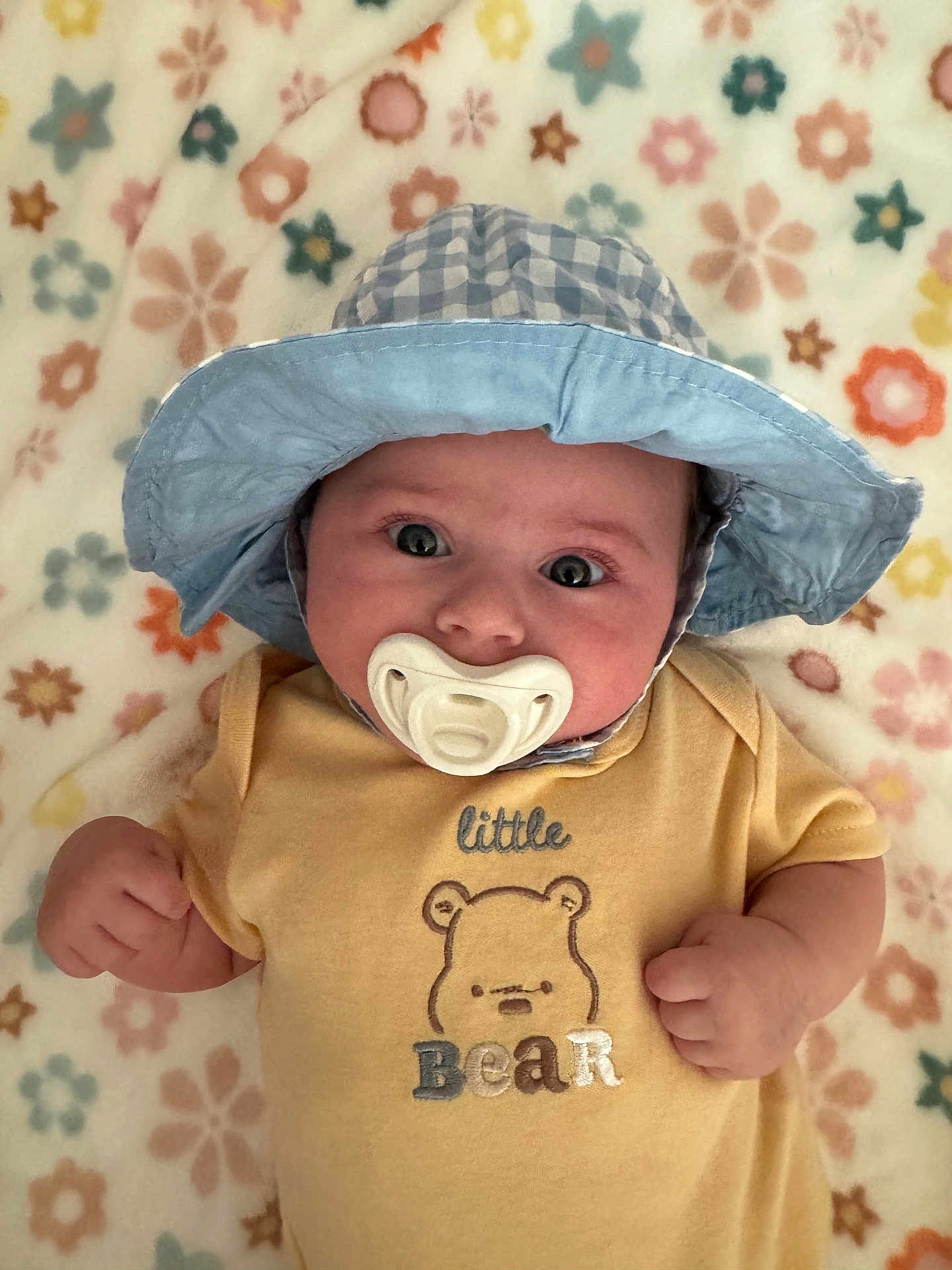Maple is registered to the contest to win money with this photo: baby, pacifier, hat, blue_hat, yellow_onesie, bear_design, flower_pattern, blanket, infant, cute, lying_down, clothing, child, portrait, indoors, soft_texture, hand, face, wide_eyes, headwear