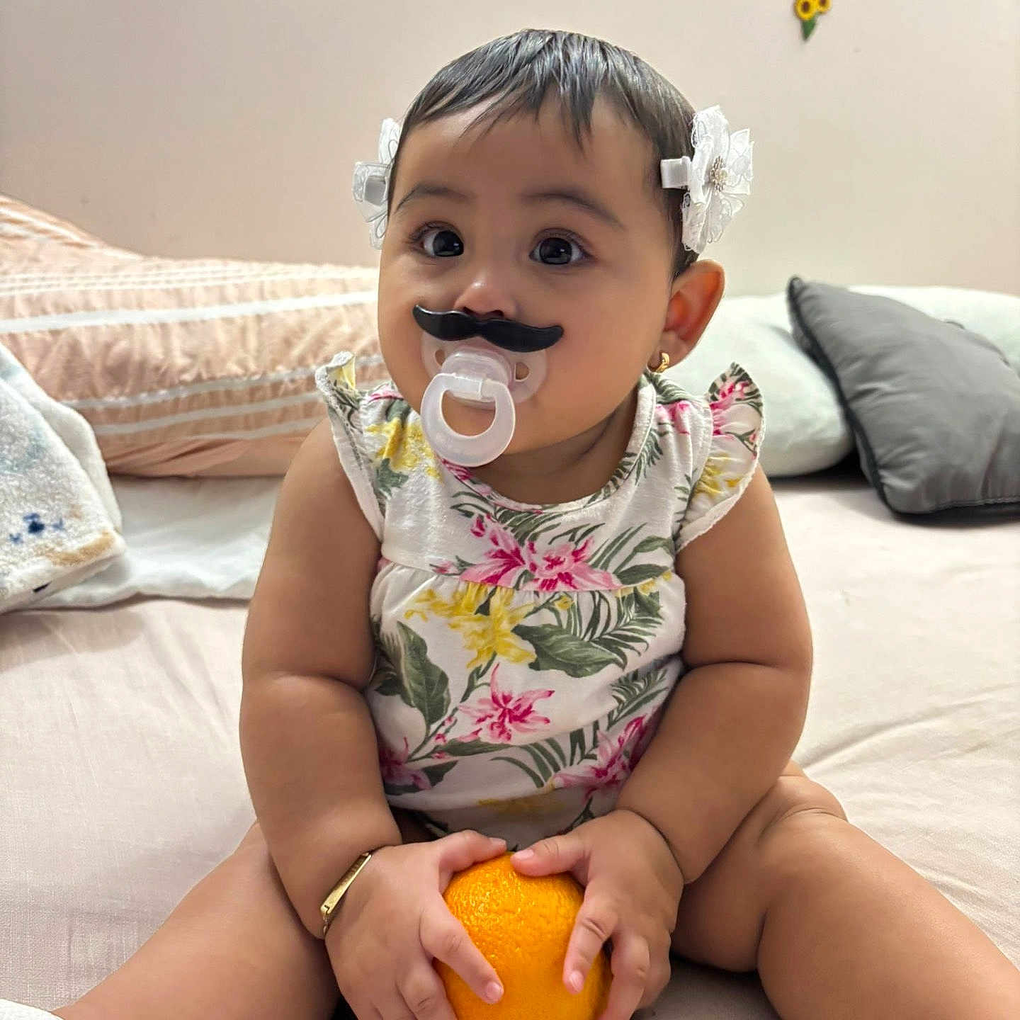 Gianella García is registered to the contest to win money with this photo: baby, bed, bracelet, child, chubby, cute, decor, flower_pattern, hair_bows, indoor, infant, mustache, orange, pacifier, pillow, playful, sitting, smile, toddler, white_shoes